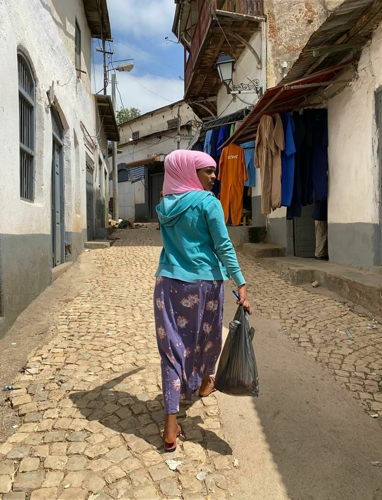A woman draped in a vibrant headscarf and flowing shawl walks through a narrow passage formed by the weathered red-brown stone walls of Harar’s Jugol, sunlight dappled across the ancient masonry.