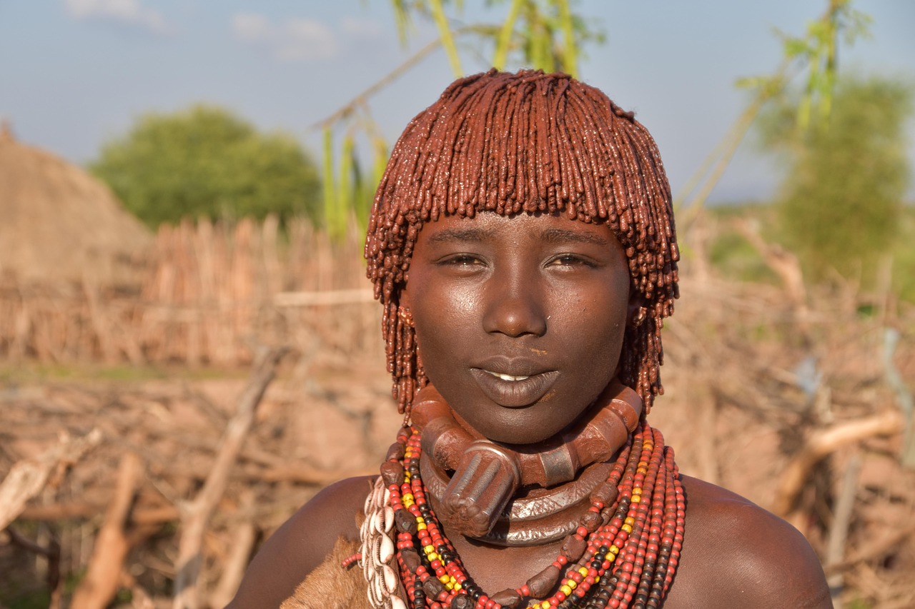 A Hamar woman stands on red earth in the Omo Valley, her hair braided and coated with red ochre, multiple metal and glass-bead necklaces around her neck, a cowrie-studded leather skirt at her waist, and stacked metal bracelets on her arms, with a thatched hut and acacia trees in soft focus behind her.